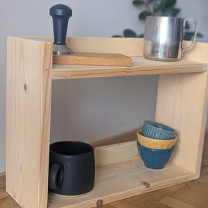 K&ouml;nnte beinhalten: Ein zweistufiges, helles Holzregal. Es enth&auml;lt einen dunkelblauen Tamper, ein kleines Holzbrett, einen Edelstahlbecher, eine schwarze Tasse und drei kleine, bunte Schalen. Das Regal steht vor einer neutralen Wand.