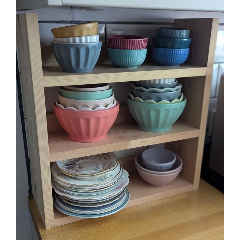 May include: A wooden shelf unit with multiple shelves, holding a variety of bowls and plates. The bowls are in various colours, including blue, green, and red. The plates are white with a floral design.