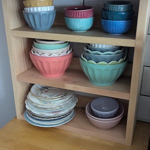 May include: A wooden shelf unit with three shelves, holding a collection of colourful ceramic bowls. The bowls are stacked and arranged in a variety of sizes and shapes. The shelves are made of natural wood and have a rustic look.