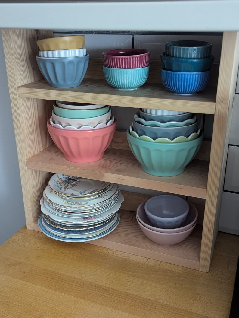 May include: A wooden shelf unit with three shelves, holding a collection of colourful ceramic bowls. The bowls are stacked and arranged in a variety of sizes and shapes. The shelves are made of natural wood and have a rustic look.