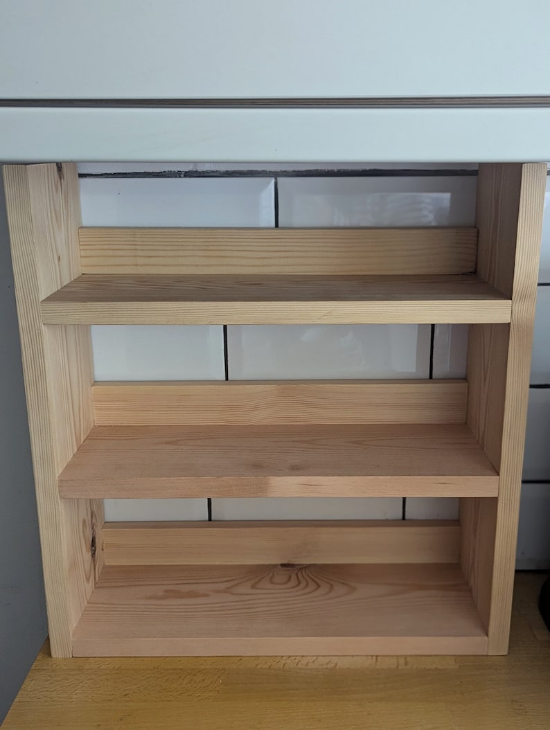 May include: A wooden shelf unit with three shelves, made of unfinished pine wood. The shelves are mounted against a white tiled wall.