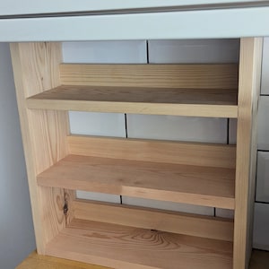 May include: A natural wood shelf unit with three shelves. The shelves are made of unfinished wood and have a light brown colour.