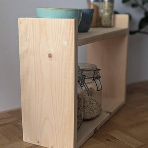 K&ouml;nnte beinhalten: Ein zweist&ouml;ckiges Holzregal in hellem Farbton. Das obere Regal enth&auml;lt zwei Schalen und zwei Gl&auml;ser mit Metallverschl&uuml;ssen. Das untere Regal enth&auml;lt ein Glas mit einem Metallverschluss. Das Regal ist aus unbehandeltem Holz gefertigt.