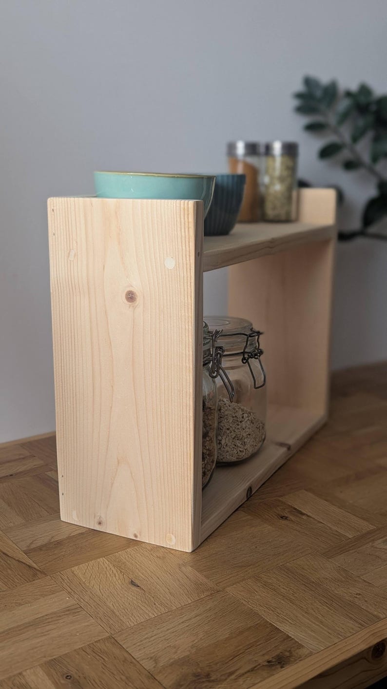 K&ouml;nnte beinhalten: Ein zweist&ouml;ckiges Holzregal in hellem Farbton. Das obere Regal enth&auml;lt zwei Schalen und zwei Gl&auml;ser mit Metallverschl&uuml;ssen. Das untere Regal enth&auml;lt ein Glas mit einem Metallverschluss. Das Regal ist aus unbehandeltem Holz gefertigt.