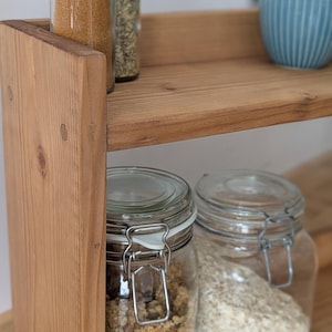 K&ouml;nnte beinhalten: Ein zweist&ouml;ckiges Gew&uuml;rzregal aus Holz mit Gl&auml;sern, die mit Gew&uuml;rzen und Getreide gef&uuml;llt sind. Das obere Regal enth&auml;lt zwei Gew&uuml;rzgl&auml;ser mit Metalldeckeln und eine kleine blaue Sch&uuml;ssel. Das untere Regal hat zwei gro&szlig;e Gl&auml;ser mit Metallverschl&uuml;ssen.