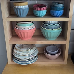 May include: A wooden shelf unit with three shelves, holding a variety of bowls and plates. The top shelf has three bowls, one yellow, one red, and one blue. The middle shelf has six bowls, two green, two blue, and two pink. The bottom shelf has a stack of plates with floral designs and two bowls, one purple and one pink.