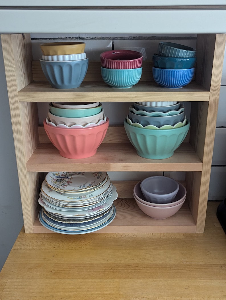 May include: A wooden shelf unit with three shelves, holding a variety of bowls and plates. The top shelf has three bowls, one yellow, one red, and one blue. The middle shelf has six bowls, two green, two blue, and two pink. The bottom shelf has a stack of plates with floral designs and two bowls, one purple and one pink.