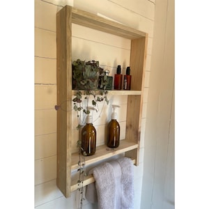 May include: A wooden bathroom shelf with two shelves and a towel rail. The shelf is made of natural wood and has a rustic look. There are two bottles of liquid soap, a plant, and two bottles of lotion on the shelves.