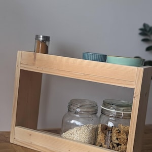 K&ouml;nnte beinhalten: Ein zweist&ouml;ckiges Gew&uuml;rzregal aus Holz. Auf der oberen Ablage befinden sich ein kleines Gew&uuml;rzglas und zwei kleine Schalen. Die untere Ablage enth&auml;lt zwei gro&szlig;e Gl&auml;ser mit Haferflocken und M&uuml;sli. Das Regal ist aus hellem Holz gefertigt.