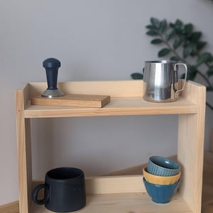 K&ouml;nnte beinhalten: Ein zweistufiges, helles Holzregal mit verschiedenen K&uuml;chenartikeln. Das obere Regal enth&auml;lt einen Kaffeestampfer, einen Edelstahlbecher und einen Holzblock. Das untere Regal hat eine schwarze Tasse und einen Stapel kleiner Schalen.