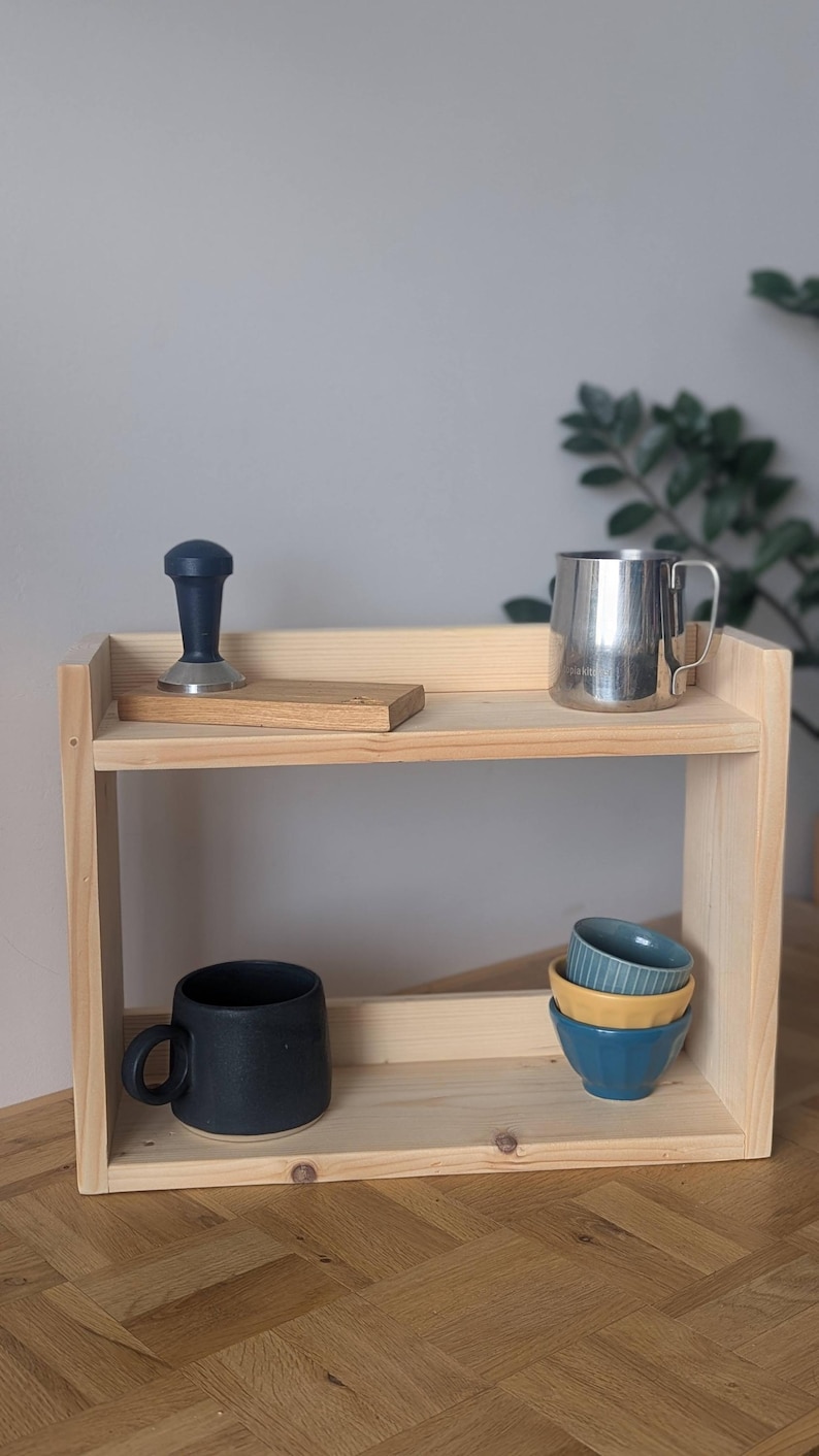 K&ouml;nnte beinhalten: Ein zweistufiges, helles Holzregal mit verschiedenen K&uuml;chenartikeln. Das obere Regal enth&auml;lt einen Kaffeestampfer, einen Edelstahlbecher und einen Holzblock. Das untere Regal hat eine schwarze Tasse und einen Stapel kleiner Schalen.