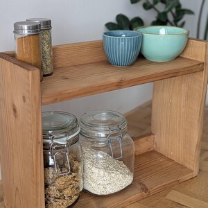 K&ouml;nnte beinhalten: Ein zweistufiges Holzregal mit verschiedenen K&uuml;chenartikeln. Das obere Regal enth&auml;lt zwei kleine Schalen, eine blaue mit wei&szlig;en Streifen und eine hellgr&uuml;ne, sowie zwei Gew&uuml;rzgl&auml;ser. Das untere Regal hat zwei Gl&auml;ser mit Metallverschl&uuml;ssen, gef&uuml;llt mit M&uuml;sli und Haferflocken.