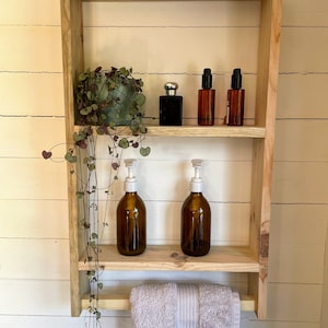 May include: A wooden bathroom shelf with two shelves and a towel rail. The shelf is holding a potted plant, two bottles of liquid soap, and three bottles of toiletries. The shelf is mounted on a white wall.