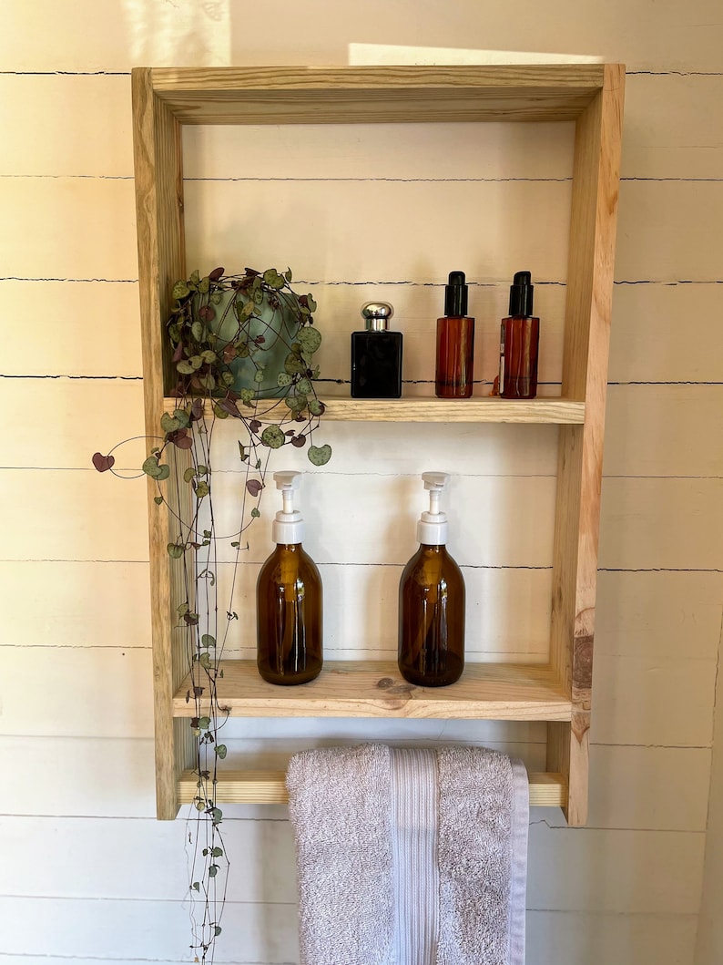 May include: A wooden bathroom shelf with two shelves and a towel rail. The shelf is holding a potted plant, two bottles of liquid soap, and three bottles of toiletries. The shelf is mounted on a white wall.