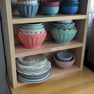 May include: A wooden shelf unit with three shelves, holding a variety of bowls and plates. The top shelf has a stack of blue bowls, a stack of teal bowls, and a stack of red bowls. The middle shelf has a stack of green bowls and a stack of blue bowls. The bottom shelf has a stack of vintage plates with floral designs.