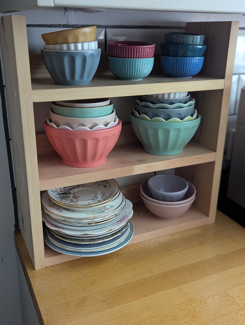 May include: A wooden shelf unit with three shelves, holding a variety of bowls and plates. The top shelf has a stack of blue bowls, a stack of teal bowls, and a stack of red bowls. The middle shelf has a stack of green bowls and a stack of blue bowls. The bottom shelf has a stack of vintage plates with floral designs.