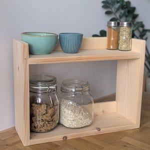 K&ouml;nnte beinhalten: Ein zweistufiges, helles Holzregal mit verschiedenen K&uuml;chenartikeln. Das obere Regal zeigt zwei kleine Schalen, eine t&uuml;rkisfarbene und eine blaue, sowie zwei Gew&uuml;rzgl&auml;ser. Das untere Regal hat zwei Gl&auml;ser mit Getreide. Eine funktionale und dekorative Aufbewahrungsl&ouml;sung.