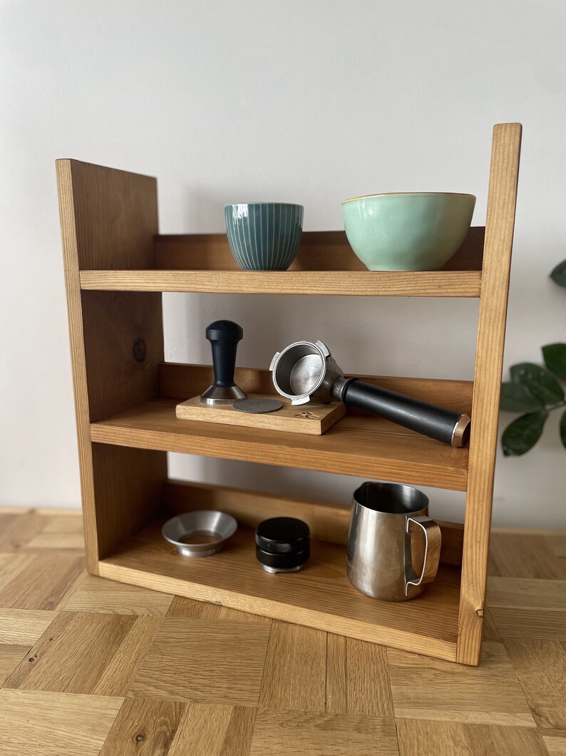 May include: A wooden shelf unit with three shelves, holding various coffee brewing tools and accessories. The top shelf has two bowls, one green and one blue. The middle shelf has a tamper, a portafilter, and a wooden base. The bottom shelf has a funnel, a tamper, and a stainless steel jug.