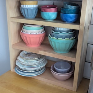 May include: A wooden shelf unit with three shelves, holding a collection of bowls and plates. The bowls are in various colours, including blue, green, pink, and red. The plates are white with gold trim.