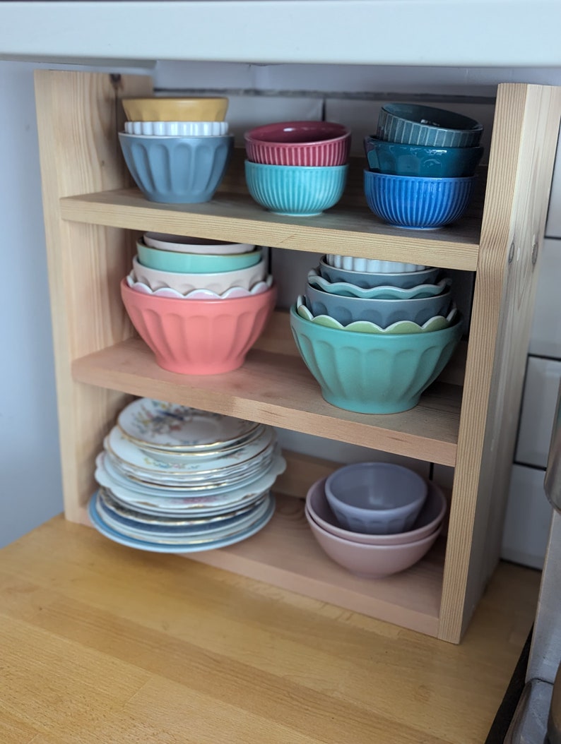 May include: A wooden shelf unit with three shelves, holding a collection of bowls and plates. The bowls are in various colours, including blue, green, pink, and red. The plates are white with gold trim.