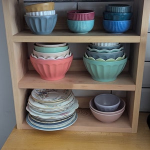 May include: A wooden shelf unit with three shelves, holding a variety of bowls and plates. The top shelf has a stack of blue bowls, a stack of teal bowls, and a stack of pink bowls. The middle shelf has a stack of green bowls, a stack of blue bowls, and a stack of teal bowls. The bottom shelf has a stack of vintage plates and a stack of pink bowls.