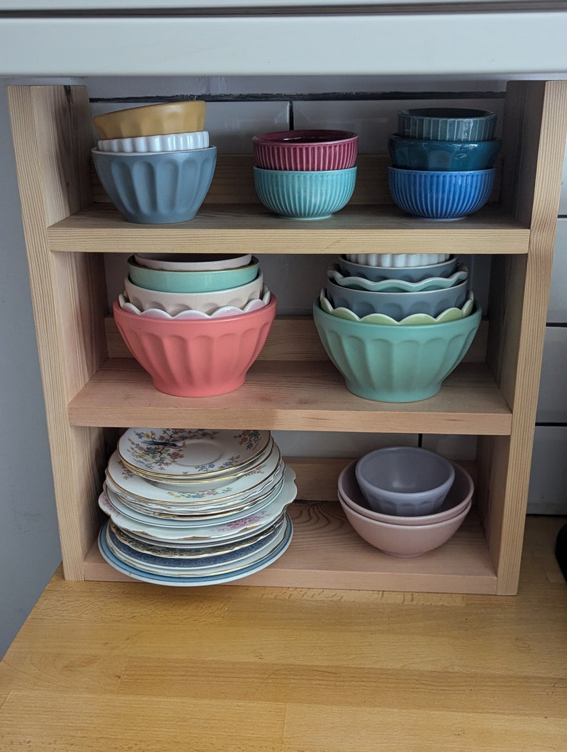 May include: A wooden shelf unit with three shelves, holding a variety of bowls and plates. The top shelf has a stack of blue bowls, a stack of teal bowls, and a stack of pink bowls. The middle shelf has a stack of green bowls, a stack of blue bowls, and a stack of teal bowls. The bottom shelf has a stack of vintage plates and a stack of pink bowls.