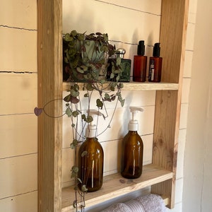 May include: A wooden shelf with two shelves and a towel rail. The shelf is made of natural wood and has a rustic look. There are two bottles of liquid soap, a plant, and two bottles of perfume on the shelves.