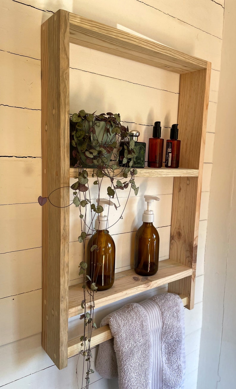 May include: A wooden shelf with two shelves and a towel rail. The shelf is made of natural wood and has a rustic look. There are two bottles of liquid soap, a plant, and two bottles of perfume on the shelves.