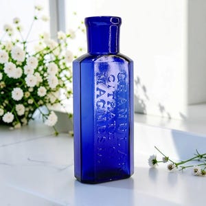 May include: A rectangular cobalt blue glass bottle with a narrow neck and a flared rim. The front face features embossed text that reads GRANULAR CITRATE OF MAGNESIA. The bottle has a smooth, reflective surface and a distinct, vintage-style shape.