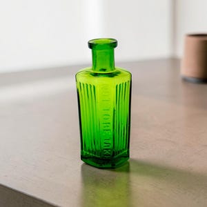 May include: A small, vibrant green glass bottle with a rectangular shape and vertical ribbed sides. The front of the bottle features embossed text that reads NOT TO BE TAKEN in capital letters. It has a narrow neck and a rounded, flared rim.