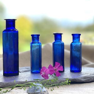 Op de afbeelding: Vier kobaltblauwe glazen flessen in verschillende maten, gerangschikt op een leisteen oppervlak. De flessen hebben een vintage uitstraling en zijn perfect om bloemen of andere decoratieve items te presenteren.