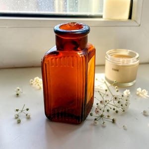 May include: A small, rectangular amber glass bottle with a thick, rounded rim and vertical ribbed detailing along the sides. The bottle features a narrow neck and a flat base, constructed from translucent brown glass that catches the light.