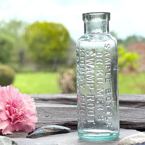 May include: A vintage clear glass sample bottle with embossed text. The bottle is next to a pink carnation flower. The text on the bottle reads "SAMPLE BOTTLE DR. KILMER'S SWAMP-ROOT KIDNEY CURE". The background is a blurred garden.