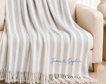 May include: A soft, woven throw blanket featuring vertical stripes in alternating shades of white and light gray. The blanket has a fringed edge at the bottom and is embroidered with the blue cursive text James & Sophia in the lower corner.