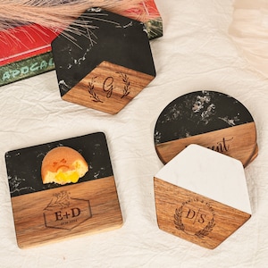 May include: Four black and white marble coasters with wooden bases. Each coaster features a different design with a monogram and a floral wreath. The coasters are ideal for protecting surfaces from heat and scratches.