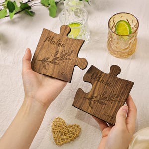 Puede incluir: Dos posavasos de madera con forma de pieza de rompecabezas, grabados con los nombres "Mateo" y "Olivia". Cada posavasos presenta un diseño floral y las palabras "his" y "hers".