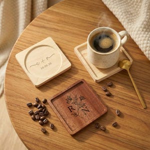 May include: Two square wooden coasters, one light maple with engraved text C + N and 10.06.26, one dark walnut with a floral design and letters R N. A speckled ceramic mug with coffee sits on a coaster, accompanied by a gold spoon and coffee beans.