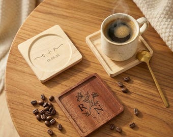 May include: Two square wooden coasters, one light maple with engraved text C + N and 10.06.26, one dark walnut with a floral design and letters R N. A speckled ceramic mug with coffee sits on a coaster, accompanied by a gold spoon and coffee beans.