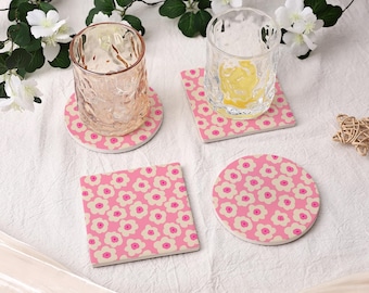 May include: Set of four drink coasters. Two square and two round coasters with a pink background and a repeating pattern of white and pink flowers. Two clear drinking glasses are on top of the coasters.