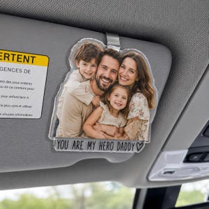 May include: A clear acrylic car visor ornament with a family photo. The image shows a smiling family of four. Text at the bottom reads "YOU ARE MY HERO DADDY" with a heart symbol. The ornament is suspended from a silver clip.