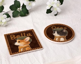 May include: Two ceramic coasters, one square and one round, featuring illustrations of pets. The square coaster has a corgi wearing a cap and scarf, with the name "Bella". The round coaster has a cat wearing a cap and scarf, with the name "Oreo".