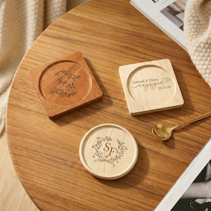 May include: Three wooden coasters with engraved designs on a wooden table. One square coaster has the initials "K M" within a floral wreath. Another square coaster reads "Samuel & Chloe engaged 05.20.26". A round coaster has "SF" in a wreath.