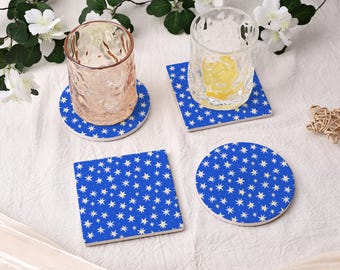 May include: Four ceramic coasters with a blue background and a white star pattern. Two coasters are square, and two are round. Two clear drinking glasses sit on top of two of the coasters. The coasters are on a white surface.