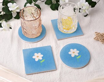 May include: A collection of drink coasters. Two round and two square coasters are light blue with a white daisy design. Two textured glass tumblers sit on the coasters. The background includes white flowers and a white tablecloth.