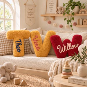 May include: Three plush, textured letter-shaped cushions in mustard yellow and deep red. The letter T is embroidered with the name Theo in blue, the letter V with Vera in pink, and the letter W with Willow in white, all in a cursive script font.