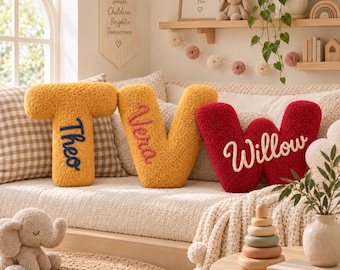 May include: Three plush, textured letter-shaped pillows in mustard yellow and deep red. The letter T is embroidered with the name Theo in blue, the letter V with Vera in pink, and the letter W with Willow in white, all in a cursive script font.