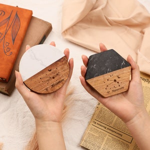 May include: Two coasters, one round and one hexagonal, are held in hands. The round coaster has a white marble top and a wooden bottom with the name "Hermosa" and a constellation design. The hexagonal coaster has a black marble top and a wooden bottom with the name "Maureen" and a constellation design.