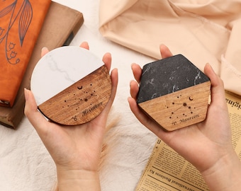 May include: Two coasters, one round and one hexagonal, are held in hands. The round coaster has a white marble top and a wooden bottom with the name "Hermosa" and a constellation design. The hexagonal coaster has a black marble top and a wooden bottom with the name "Maureen" and a constellation design.