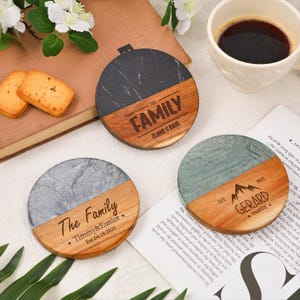 Puede incluir: Tres posavasos personalizados con una combinación de madera y piedra. Cada posavasos presenta un nombre familiar y una fecha, con el texto "The Family" o "Gerard Family" grabado en la madera. Una taza de café y galletas están en el fondo.