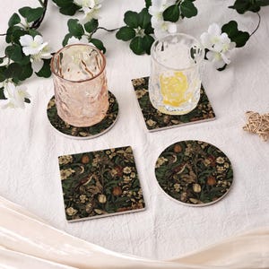 May include: Four ceramic coasters featuring a floral and bird design in green, brown, and cream. Two clear drinking glasses are placed on two of the coasters. One coaster is round, one is square, and two are round.